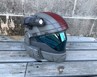 May include: A gray and red helmet with a visor and a flashlight on top. The helmet has a textured surface and a red stripe on the side.