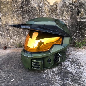Könnte beinhalten: Ein grüner und schwarzer Helm mit einem goldenen Visier, das die Umgebung reflektiert. Der Helm hat ein Visier und einen Kinnschutz. Er besteht aus einem robusten Material und ist wahrscheinlich für Cosplay oder Ausstellungszwecke gedacht.