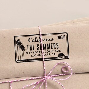 May include: A stack of kraft paper envelopes tied with pink and white twine. Each envelope is stamped with a black address label that reads "California, The Summers, 2367 Pacific Coast Ave, Los Angeles, CA". A wooden stamp and a spool of twine are also visible.