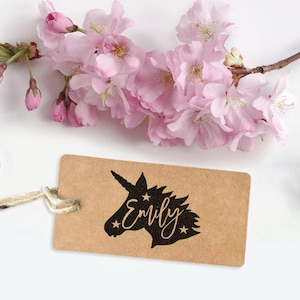 May include: A brown paper tag with a black silhouette of a unicorn's head and the name "Emily" written in a cursive font. The tag is tied with a white string and surrounded by pink cherry blossoms.