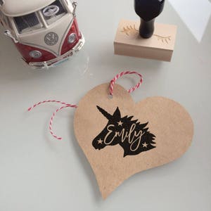 May include: A brown heart-shaped tag with a black silhouette of a unicorn with the name "Emily" written in cursive. The tag is tied with a red and white string.