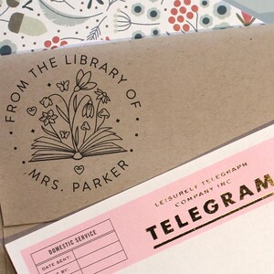 May include: A brown paper envelope featuring a black ink stamp with an open book, flowers, butterflies, and the text FROM THE LIBRARY OF MRS. PARKER, positioned next to a pink telegram form with gold foil text reading LEISURELY TELEGRAPH COMPANY INC.