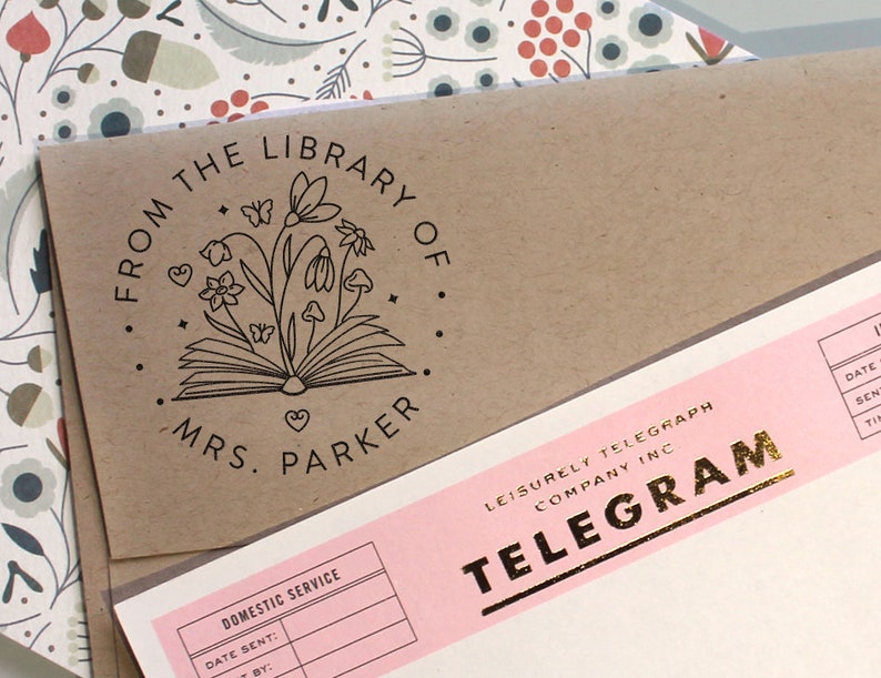 May include: A brown paper envelope featuring a black ink stamp with an open book, flowers, butterflies, and the text FROM THE LIBRARY OF MRS. PARKER, positioned next to a pink telegram form with gold foil text reading LEISURELY TELEGRAPH COMPANY INC.