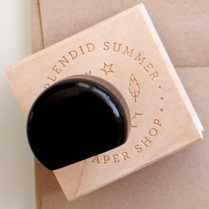 May include: A wooden stamp with a black handle. The stamp features the text "Splendid Summer Paper Shop" in a circle around a star and a feather design. The stamp is mounted on a wooden block.