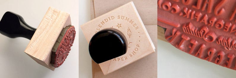 May include: A wooden stamp with a black handle. The stamp features the text "Splendid Summer Paper Shop" in a circle around a star and a feather design. The stamp is mounted on a wooden block.