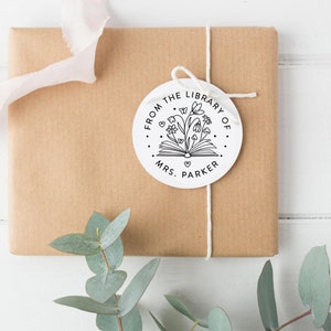 May include: A circular white gift tag featuring a black line art illustration of an open book with flowers, butterflies, and small hearts emerging from the pages. The text reads FROM THE LIBRARY OF MRS. PARKER in a clean, sans-serif font.