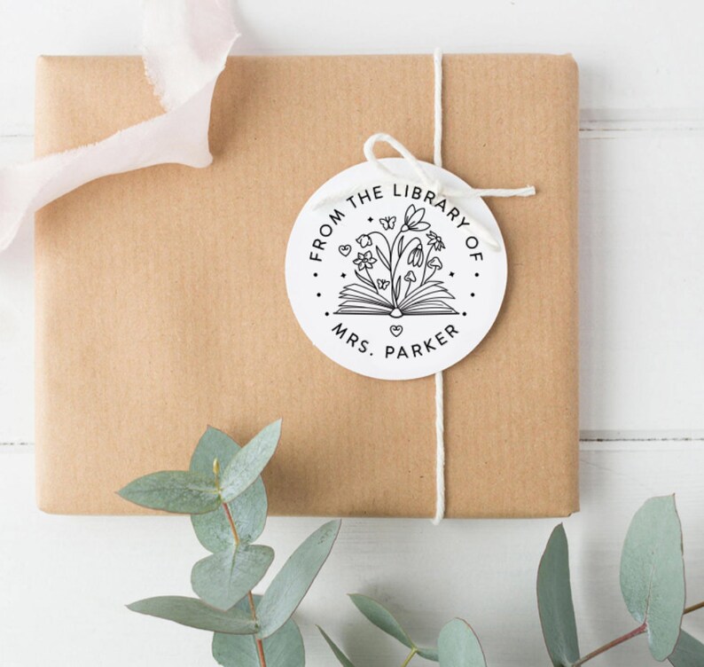 May include: A circular white gift tag featuring a black line art illustration of an open book with flowers, butterflies, and small hearts emerging from the pages. The text reads FROM THE LIBRARY OF MRS. PARKER in a clean, sans-serif font.