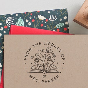 May include: A circular rubber stamp impression on tan paper featuring an open book with flowers, mushrooms, and butterflies emerging from the pages. The text reads FROM THE LIBRARY OF MRS. PARKER, surrounded by a dotted border and small stars.