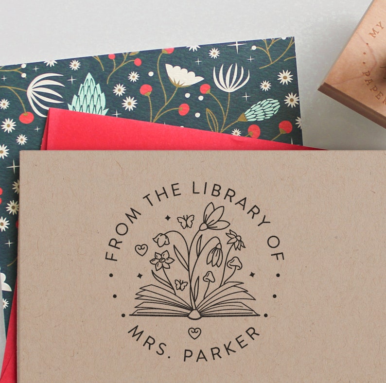 May include: A circular rubber stamp impression on tan paper featuring an open book with flowers, mushrooms, and butterflies emerging from the pages. The text reads FROM THE LIBRARY OF MRS. PARKER, surrounded by a dotted border and small stars.