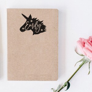 May include: A brown notebook with a black unicorn silhouette and the name "Emily" written in cursive. The notebook is on a white surface with a pink rose.