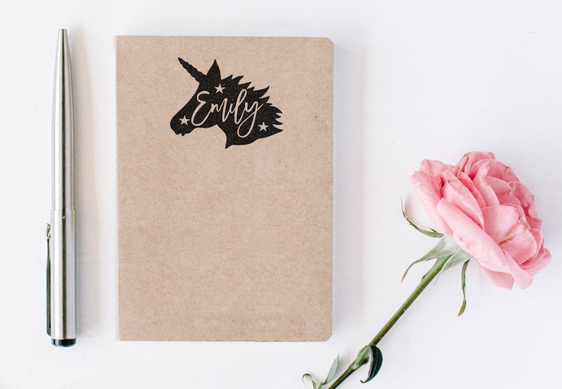 May include: A brown notebook with a black unicorn silhouette and the name "Emily" written in cursive. The notebook is on a white surface with a pink rose.