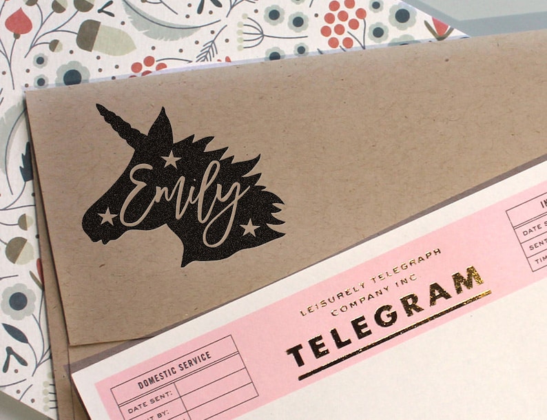 May include: A brown paper envelope with a black unicorn silhouette and the name "Emily" printed on it. The envelope is partially covering a pink telegram form with gold lettering.