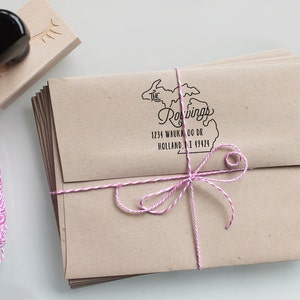 May include: A stack of brown envelopes tied with pink and white twine. The envelopes are addressed with a rubber stamp that says "The Roavings" and includes an outline of the state of Michigan. The address is 1234 Waukoo Dr, Holland, MI 49424.