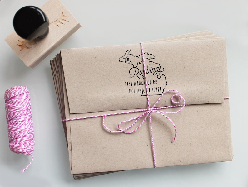 May include: A stack of brown envelopes tied with pink and white twine. The envelopes are addressed with a rubber stamp that says "The Roavings" and includes an outline of the state of Michigan. The address is 1234 Waukoo Dr, Holland, MI 49424.