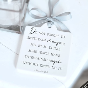 May include: White ceramic ornament with a black quote: "Do not forget to entertain strangers, for by so doing, some people have entertained angels without knowing it. - Hebrews 13:2". The ornament has a gray ribbon attached.