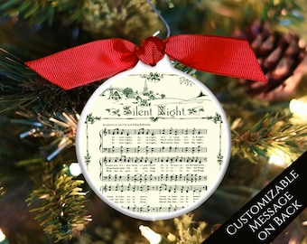 Könnte beinhalten: Weißer Keramik-Weihnachtsbaumschmuck mit einer Vintage-Illustration eines Winterdorfes und der Noten für das Lied "Stille Nacht". Der Anhänger hat ein rotes Band und einen silbernen Haken. Die Rückseite des Anhängers ist mit einer Nachricht personalisierbar.