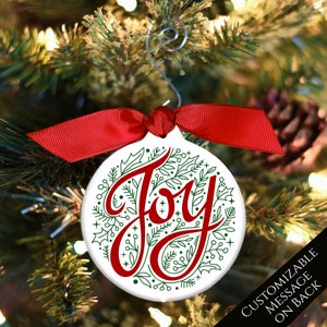 May include: A white ceramic Christmas ornament with a red ribbon. The ornament features a green and white floral design with the word "Joy" in red lettering.