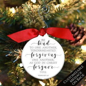 May include: White round Christmas ornament with black text that reads "BE kind TO ONE ANOTHER, TENDERHEARTED, forgiving ONE ANOTHER, AS GOD IN CHRIST forgave YOU. - EPHESIANS 4:32". The ornament is hanging from a red ribbon on a Christmas tree.