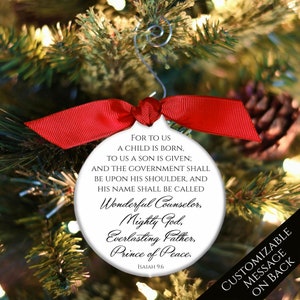 May include: White round Christmas ornament with a red ribbon. The ornament features a black and white text quote from Isaiah 9:6, "For to us a child is born, to us a son is given; and the government shall be upon his shoulder, and his name shall be called Wonderful Counselor, Mighty God, Everlasting Father, Prince of Peace."