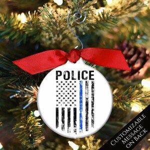 May include: A white round Christmas ornament with a blue line American flag design and the word "POLICE" printed in black at the top. The ornament is hanging from a red ribbon on a Christmas tree.