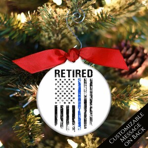 May include: A white round Christmas ornament with a blue line American flag design and the word "RETIRED" printed in black. The ornament has a red ribbon and a silver hanger.