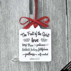 May include: A white, square ornament with a red ribbon and bow. The ornament features the text "The Fruit of the Spirit" followed by "love, joy, peace, patience, kindness, goodness, faithfulness, gentleness, self-control" and "Galatians 5:22-23".