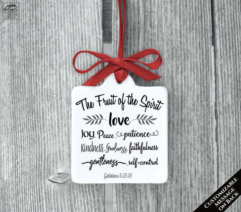 May include: A white, square ornament with a red ribbon and bow. The ornament features the text "The Fruit of the Spirit" followed by "love, joy, peace, patience, kindness, goodness, faithfulness, gentleness, self-control" and "Galatians 5:22-23".