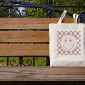 May include: A white tote bag with a pink checkered pattern and a pink smiley face with lightning bolts for eyes. The bag is sitting on a wooden bench.