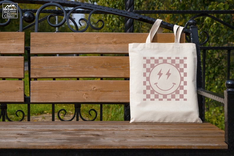 May include: A white tote bag with a pink checkered pattern and a pink smiley face with lightning bolts for eyes. The bag is sitting on a wooden bench.