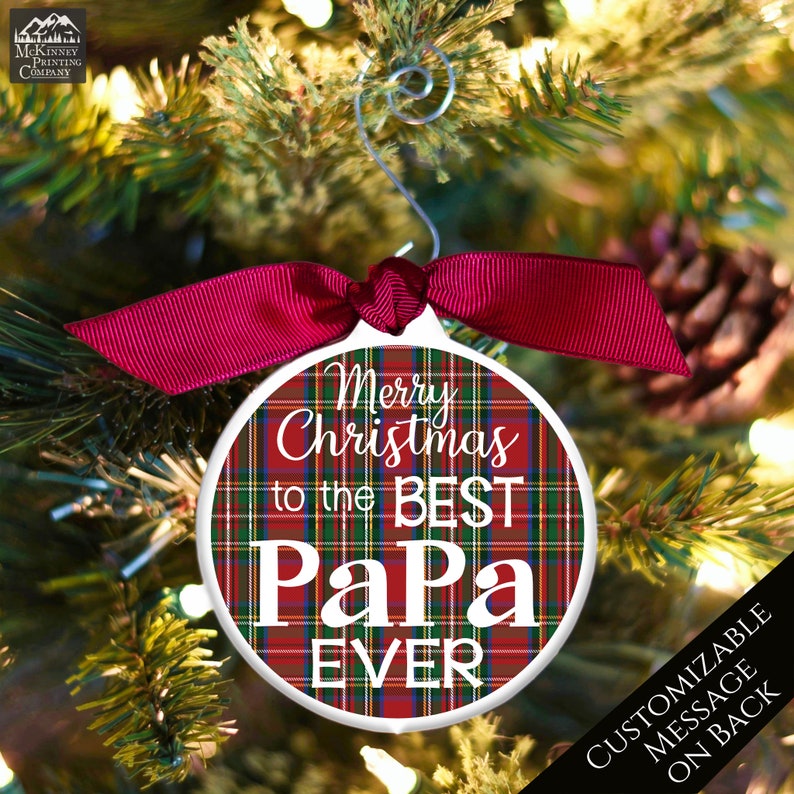 May include: Red and green plaid Christmas ornament with white text that reads "Merry Christmas to the Best Papa Ever". The ornament is hanging on a Christmas tree with red ribbon.