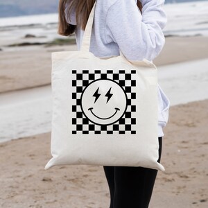 May include: A white tote bag with black handles features a black and white chequered pattern with a black smiley face with lightning bolts for eyes. The bag is being held by a person wearing a grey hoodie and black leggings.