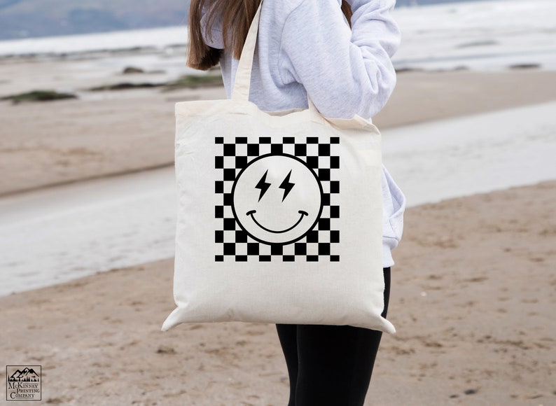 May include: A white tote bag with black handles features a black and white chequered pattern with a black smiley face with lightning bolts for eyes. The bag is being held by a person wearing a grey hoodie and black leggings.