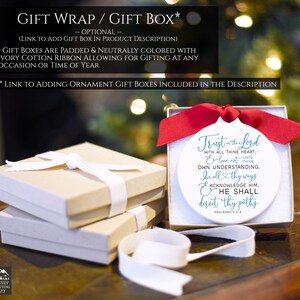 May include: A white ornament with a red ribbon, featuring the text "Trust in the Lord." The ornament is in a gift box with a white ribbon. Additional gift boxes are stacked nearby, tied with ivory ribbons. Text at the top reads "Gift Wrap / Gift Box."