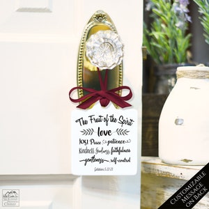 May include: A white door hanger with a burgundy ribbon and a decorative door knob. The hanger features the text "The Fruit of the Spirit" with a list of virtues and the scripture reference Galatians 5:22-23.
