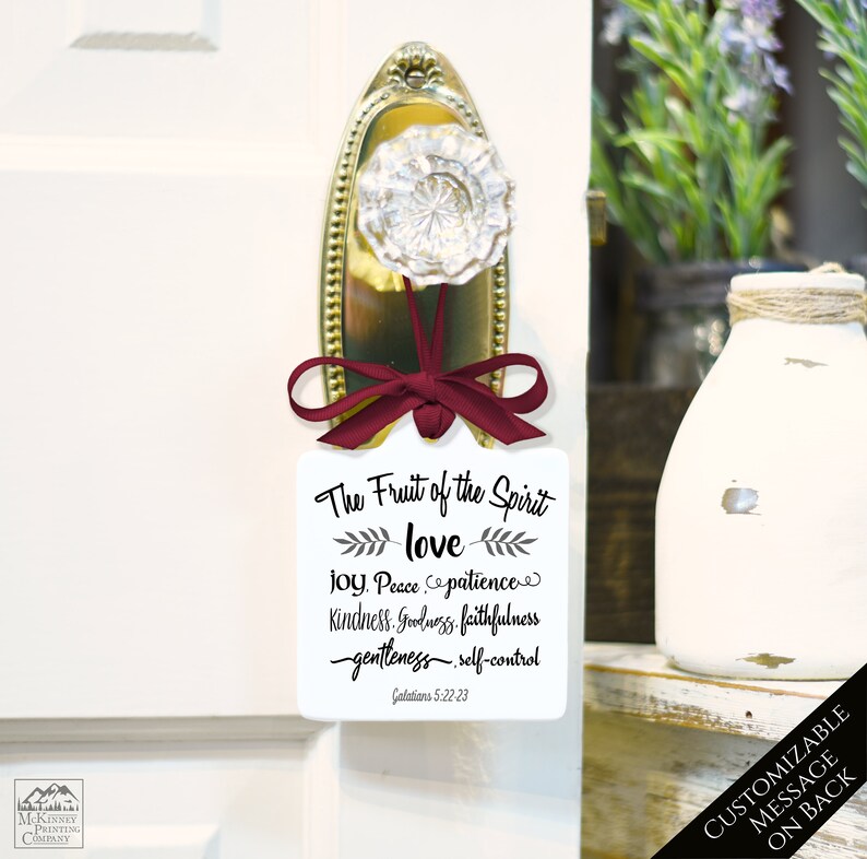 May include: A white door hanger with a burgundy ribbon and a decorative door knob. The hanger features the text "The Fruit of the Spirit" with a list of virtues and the scripture reference Galatians 5:22-23.
