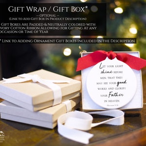 May include: A white ceramic ornament with the text "Let your light shine before men. That they may see your good works and glorify your Father in Heaven. -Matthew 5:16". The ornament is in a white gift box with a red ribbon. The gift box is sitting on a wooden surface.