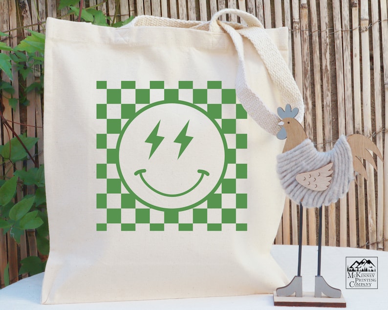 May include: A white canvas tote bag featuring a green checkered pattern and a green smiley face with lightning bolts for eyes. The text "McKinsey Printing Company" is printed in the bottom right corner of the bag.