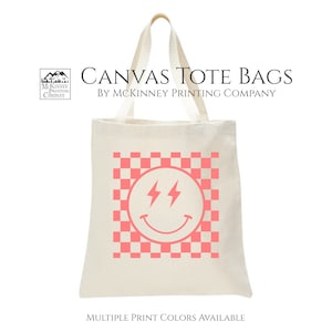 May include: A white canvas tote bag featuring a pink checkered pattern and a pink smiley face with lightning bolts for eyes. The text "CANVAS TOTE BAGS BY MCKINNEY PRINTING COMPANY" is printed at the top of the bag. The text "MULTIPLE PRINT COLORS AVAILABLE" is printed at the bottom of the bag.