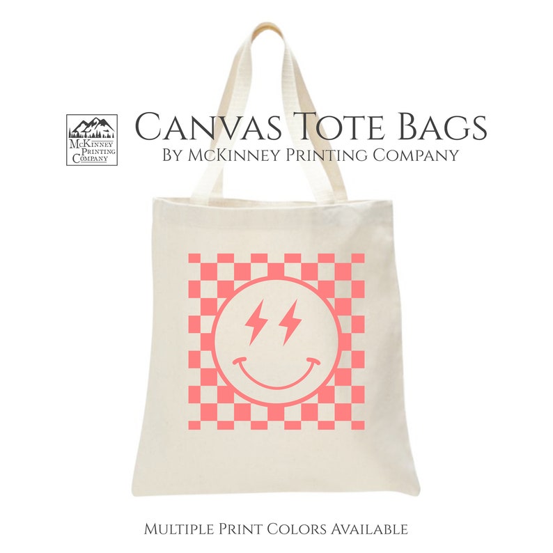 May include: A white canvas tote bag featuring a pink checkered pattern and a pink smiley face with lightning bolts for eyes. The text "CANVAS TOTE BAGS BY MCKINNEY PRINTING COMPANY" is printed at the top of the bag. The text "MULTIPLE PRINT COLORS AVAILABLE" is printed at the bottom of the bag.