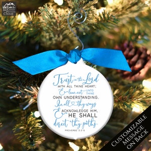 May include: White Christmas ornament with a blue ribbon and the text "Trust in the Lord" with a bible verse. The ornament is hanging on a Christmas tree. The back of the ornament is customizable.