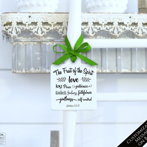 May include: A white, rectangular tag with the text "The Fruit of the Spirit" and related words in black script. A green ribbon is tied at the top. The tag is hanging on a white pole. The background includes a decorative shelf.