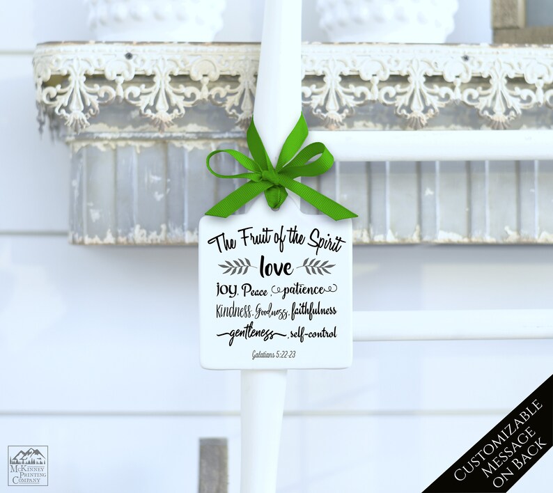 May include: A white, rectangular tag with the text "The Fruit of the Spirit" and related words in black script. A green ribbon is tied at the top. The tag is hanging on a white pole. The background includes a decorative shelf.