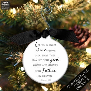 May include: White round Christmas ornament with black text that reads "Let your light shine before men, that they may see your good works and glorify your Father in heaven. -Matthew 5:16". The ornament is hanging on a Christmas tree with lights.
