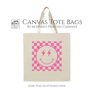 May include: A white canvas tote bag with a pink checkered pattern and a pink smiley face with lightning bolt eyes. The text "CANVAS TOTE BAGS BY MCKINNEY PRINTING COMPANY" is printed above the design. The text "LYING FLAT (FLATTENED) VIEW" is printed below the design.