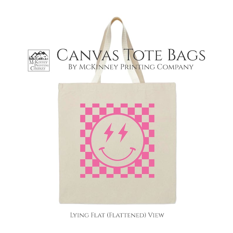 May include: A white canvas tote bag with a pink checkered pattern and a pink smiley face with lightning bolt eyes. The text "CANVAS TOTE BAGS BY MCKINNEY PRINTING COMPANY" is printed above the design. The text "LYING FLAT (FLATTENED) VIEW" is printed below the design.