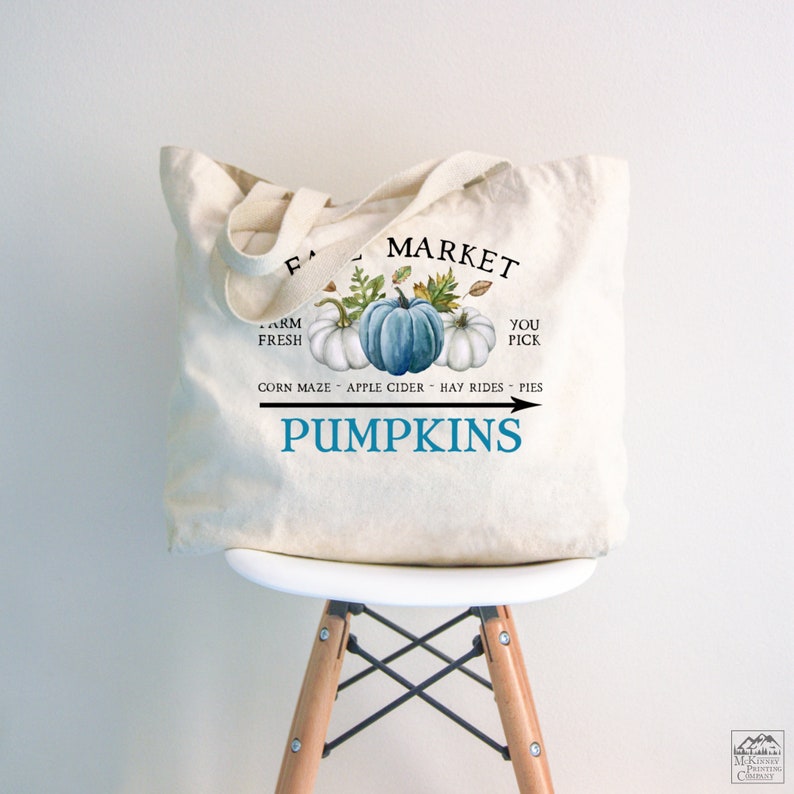 Op de afbeelding: Een witte canvas draagtas met een blauw en wit pompoenontwerp. De tekst "Fall Market" is in zwart bovenaan de tas gedrukt. De tekst "Farm Fresh" is in zwart aan de linkerkant van de tas gedrukt. De tekst "You Pick" is in zwart aan de rechterkant van de tas gedrukt. De tekst "Corn Maze - Apple Cider - Hay Rides - Pies" is in zwart onder de pompoenen gedrukt. De tekst "Pumpkins" is in blauw met een pijl naar rechts gedrukt.