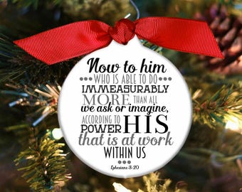 May include: White round Christmas ornament with a red ribbon. The ornament features a black and gray text design that reads "Now to him... who is able to do... immeasurably more, than all we ask or imagine, according to his power that is at work within us... Ephesians 3:20".