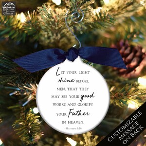 May include: White round Christmas ornament with a blue ribbon. The ornament has a black and white quote from Matthew 5:16, "Let your light shine before men, that they may see your good works and glorify your Father in heaven."