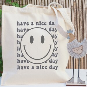 May include: A white canvas tote bag with a gray smiley face and the text "have a nice day" repeated multiple times in a retro font.