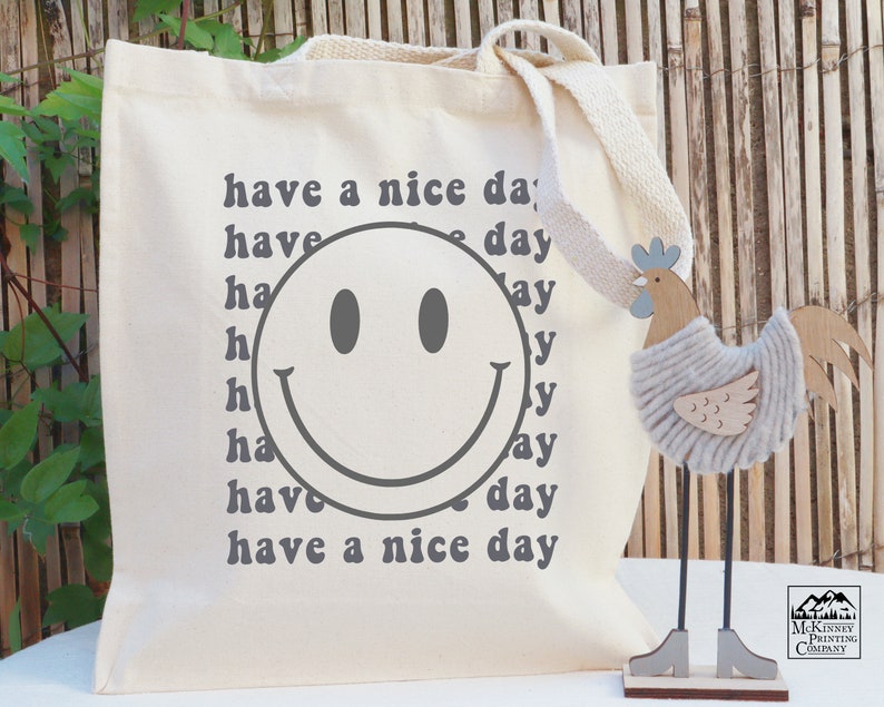 May include: A white canvas tote bag with a gray smiley face and the text "have a nice day" repeated multiple times in a retro font.